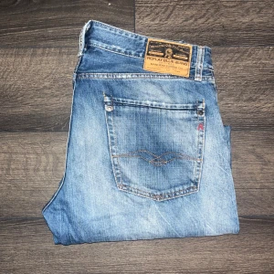 Replay jeans straight fit - Säljer ett par klassiska blå Replay jeans med snyggt slitna detaljer och raka ben. Jeansen har fem fickor, läderpatch bak och diskret broderi på bakfickan. De är tillverkade i bomull och har en normal passform med midwaist. Modell Rogob. Midja 42cm, längd 104cm. JAG SKICKAR INGA FLER BILDER PÅ NÄR JAG BÄR JEANSEN :)  S2