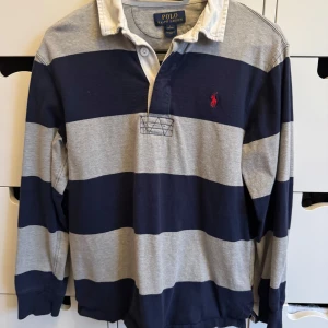Randig pikétröja från Polo Ralph Lauren (14-16 years) - Snygg långärmad pikétröja från Polo Ralph Lauren med breda marinblå och grå ränder. Klassisk krage, knäppning vid halsen och ikonisk röd logga broderad på bröstet. Perfekt för dig som gillar preppy stil och vill sticka ut lite extra.