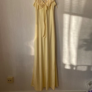 Ljusgul långklänning med volang - Supersöt långklänning i ljusgul nyans med tunna axelband, volangdetaljer vid urringningen och knyt framtill. Materiale är mjukt, tunt och lätt. 