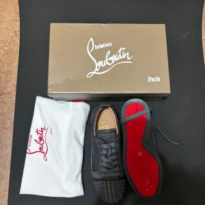 Christian Louboutin skor - Säljer ett par grå sneakers från Christian Louboutin med ikonisk röd sula och coola nitar på tån. Skorna har snörning och är tillverkade i mocka med detaljer i skinn. Perfekt för dig som vill sticka ut med en edgy och lyxig stil.