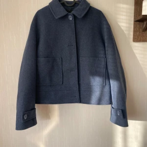 Mörkblå jacka  - Snygg mörkblå jacka med klassisk krage och stora, fyrkantiga fickor framtill. Jackan har knappar både framtill och vid ärmsluten, vilket ger en clean och stilren look. Perfekt för dig som gillar minimalistisk design och vill ha något som funkar till många outfits.