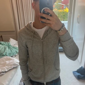 Grå hoodie med dragkedja från John Henric - Säljer en stilren ljusgrå hoodie från John Henric med dragkedja framtill och huva. Tröjan har långa ärmar, ribbade muddar och två fickor framtill. Perfekt för chill dagar eller när du vill ha en enkel och clean look.