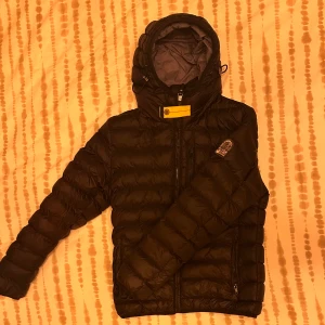 Svart dunjacka från Parajumpers - Svart dunjacka från Parajumpers med huva, gul detalj vid halsen och ikonisk PJS-patch på ärmen. Jackan är superlätt, har två dragkedjefickor och quiltad design. Perfekt för kalla dagar och har en clean, sportig look som funkar till streetwear. Pris diskuteras vid snabb affär 