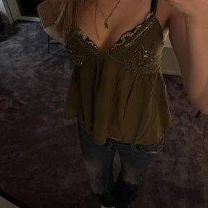 Olivgrön linnetopp med pärldetaljer - Supersöt olivgrön linnetopp från stradivarius med tunna axelband och v-ringning. Toppen har broderade pärldetaljer och paljetter upptill samt en lös, flowy passform. Svart  spetskant vid bysten ger en extra touch. Perfekt att styla med jeans för en cool look 💕