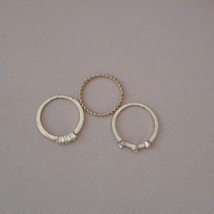 Set med tre tunna guldringar - Säljer ett set med tre tunna guldfärgade ringar. Två av ringarna har små klara stenar infattade i rad, medan den tredje har en tvinnad design utan stenar. Perfekt för att mixa och matcha på fingrarna för en snygg look.