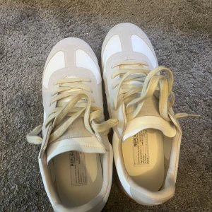 Maison Martin Margiela sneakers beige/vit - Maison Martin Margiela sneakers i beige och vitt med klassisk gummisula. Ovandel i skinn och mocka, neutrala färger och snygg retrostil. Kommer med originalkartong, dustbags och papper. Perfekta för dig som gillar minimalistisk och stilren design.