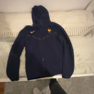 Blå Nike vindjacka Frankrike landslag - Mörkblå vindjacka från Nike med Frankrikes landslagslogga på bröstet och hel dragkedja framtill. Jackan har huva och långa ärmar, perfekt för dig som gillar sportig stil och vill visa ditt stöd för Frankrike.