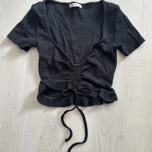 Svart topp från Zara med snörning - Svart ribbad topp från Zara med korta ärmar, djup v-ringning och snygg snörning framtill som ger en croppad look. Toppen har volangkant nedtill och är i toppskick.
