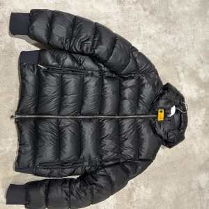 Svart pufferjacka från Parajumpers - Snygg svart pufferjacka från Parajumpers med klassisk gul detalj vid kragen. Jackan har dragkedja framtill, två sidofickor med dragkedja och ribbade muddar vid ärmslut. Perfekt för kalla dagar med sin vadderade design och höga krage. Storlek L men passar även M