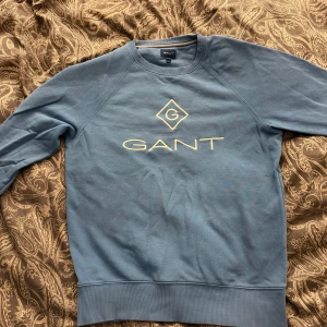 Blå sweatshirt från GANT med logga - Säljer en blå sweatshirt från GANT med broderad logga i vitt på bröstet. Tröjan har rund halsringning, Perfekt för en stilren look.