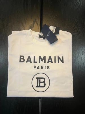 Vit Balmain Paris t-shirt - Snygg vit t-shirt från Balmain Paris med svart logotyptryck framtill. Klassisk rund halsringning och korta ärmar. Tillverkad i mjuk bomull för en skön känsla. Perfekt för dig som gillar stilrena och trendiga plagg.