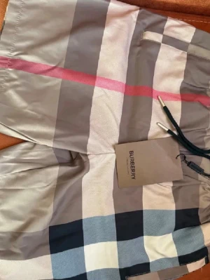 Burberry shorts, HELT NY!!! - Snygga badbyxor från Burberry med klassiskt rutmönster i grått, vitt, svart och rött. Modellen har dragsko i midjan och ficka bak. Tillverkade i ett lätt och snabbtorkande material, perfekta för stranden eller poolen.
