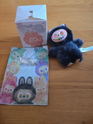 Mjukisdjur med regnbågsögon - Unikt mjukisdjur med svart fluffig päls och stora regnbågsfärgade ögon. Ansiktet har ett brett leende med färgglada detaljer och små öron på toppen. Perfekt som dekoration eller samlarobjekt för dig som gillar annorlunda gosedjur.