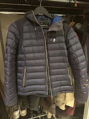 Mörkblå skidjacka  från Peak Performance - Snygg mörkblå skidjacka från Peak Performance med huva och dragkedja. Jackan har två framfickor med dragkedja och en blåmönstrad insida. Perfekt för kalla vinterdagar tack vare det värmande dunet och den sportiga designen. Storlek M men skulle säga att det är mer av en S