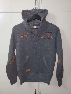 Grå hoodie med knappar och patchar - Grå hoodie med huva, bruna knappar vid halsen och broderade texter på bröstet. Har känguruficka framtill och bruna mockadetaljer på armbågarna. Snygg kontrast med orange brodyr och patch vid fickan. Perfekt för en avslappnad streetstil.