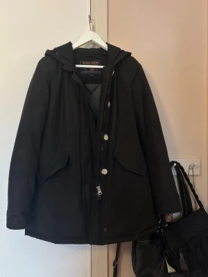 Svart dunjacka från Woolrich - Svart dunjacka från Woolrich med huva, dragkedja och knappar framtill. Jackan har två stora fickor med lock och quiltat foder i grått. Bra använt skick förutom ett mindre jack ( se bild 4)