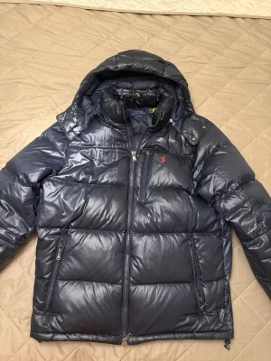 Mörkblå dunjacka från Polo Ralph Lauren - Snygg mörkblå dunjacka från Polo Ralph Lauren med avtagbar huva och dragkedja framtill. Jackan har två sidofickor med dragkedja och en bröstficka med röd logga. Perfekt för kalla vinterdagar och riktigt stilren look.