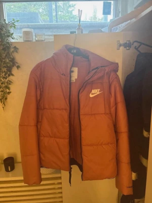 Rosa Nike pufferjacka med huva - Säljer en rosa pufferjacka från Nike med Therma-Fit-teknologi. Jackan har huva, dragkedja framtill och Nike-logga i vitt på bröstet. Perfekt för kalla dagar och riktigt skön att bära. Jackan är vadderad och har en modern, boxig passform.