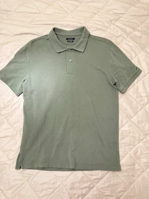 Grön pikétröja från Massimo dutti - Klassisk grön pikétröja från Massimo dutti med korta ärmar och krage. Tröjan har knappar vid halsen och är tillverkad i mjuk bomull som känns skön mot huden. Perfekt för en avslappnad och stilren look.