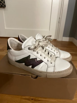 Vita sneakers med blixtdetalj Zadig & Voltaire - Säljer ett par vita sneakers från Zadig & Voltaire med metallicfärgad blixtdetalj i lila och grön på sidan. Skorna har snörning och är tillverkade i skinn. Säljer pga att de är för stora, ny pris 2900 pris kan diskuteras.💞 