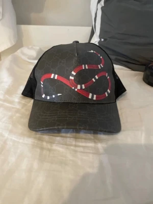Gucci keps med orm och mesh - Snygg svart keps från Gucci med ikoniskt ormmotiv i rött, vitt och svart framtill. Kepsen har diskret GG-mönster i tyget och mesh på sidorna för extra ventilation. Justerbar baktill för perfekt passform. Perfekt accessoar för en trendig streetstyle.