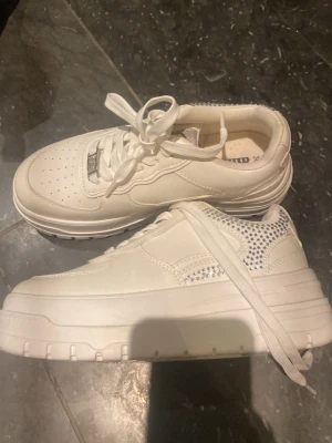Vita chunky sneakers med strassdetaljer - Säljer ett par vita chunky sneakers med platåsula och coola strassdetaljer på sidan. Skorna har perforerad tå, vita skosnören och är tillverkade i syntetmaterial. Perfekta för dig som vill sticka ut med en trendig och sportig look. Orginal pris 400kr. Storlek 36