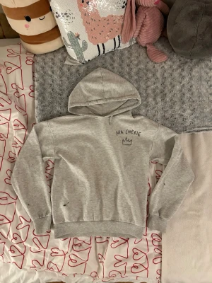 Grå hoodie med tryck från Gina tricot - Grå hoodie från Gina tricot med huva och trycket 'MA CHÉRIE' samt en krona både fram och bak. Tröjan har fläckar från grön akrylfärg. Mjuk bomullskänsla, ribbade muddar vid ärmar och midja.