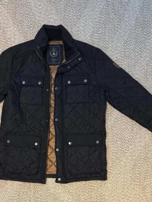Svart quiltad jacka från Marine Classic - Snygg svart quiltad jacka från Marine Classic med fyra fickor framtill och tryckknappar. Jackan har ståkrage, dragkedja och bruna detaljer på insidan. Perfekt för dig som gillar stilren och klassisk look.