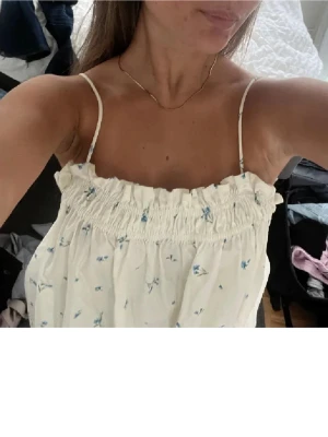 Vit blommig klänning  - Supersöt vit klänning med tunna axelband och små blå blommor. Klänningen har volangdetalj upptill och en loose passform som känns luftig och perfekt för varma dagar.