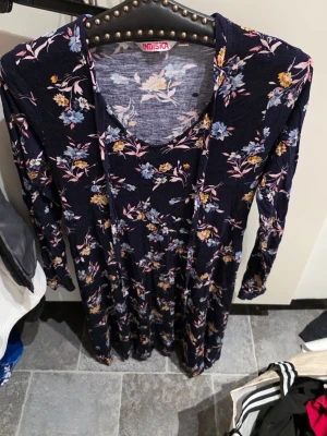Mörkblå blommig klänning Indiska - Mörkblå långärmad klänning från Indiska med blommigt mönster i blått, gult och rosa. Klänningen har en rund halsringning och är i ett mjukt, stretchigt material som ger en bekväm passform. Perfekt för dig som gillar färgglada prints och en avslappnad stil.