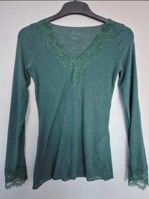Grön långärmad topp med spets - Grön långärmad topp  med v-ringning och spetsdetaljer både vid halsen och ärmsluten från Intimissimi. Perfekt för dig som gillar en enkel men ändå söt stil. Toppen har en smickrande passform och är lätt att matcha med jeans eller kjol. Den är i storlek S och i Kashmir.