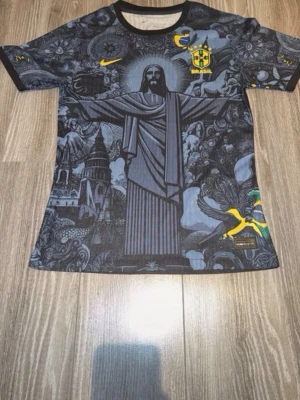 Brasilien fotbollströja svart Nike - Snygg svart Brasilien fotbollströja från Nike med unikt grafiskt tryck av Kristusstatyn och brasilianska motiv. Tröjan har gula detaljer, CBF-logga och rund hals. Perfekt för dig som vill sticka ut på planen eller heja på landslaget.