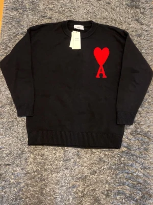 Svart stickad tröja från Ami Paris - Säljer en svart stickad tröja från Ami Paris med deras ikoniska röda hjärta och bokstaven A broderat på bröstet. Tröjan har rund hals, ribbade muddar och nederkant. Perfekt för dig som gillar stilrena plagg med en twist.