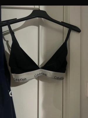 Svart Calvin Klein bralette - Svart bralette från Calvin Klein med vit resår och logga runt om. Triangelmodell utan bygel och med smala axelband. Perfekt för dig som gillar en enkel och stilren look. Materialet är mjukt och stretchigt för extra komfort.