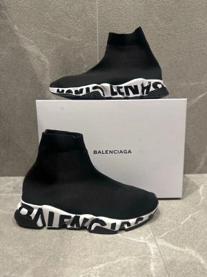 Balenciaga Speed Trainer svarta sneakers - Svarta Balenciaga Speed Trainer sneakers med stickad strumpdesign och vit chunky sula. Ena paret har stor logotyp i svart och vitt längs sulan, medan det andra har en mer diskret vit sula. Materialet är stretchig textil som sitter tight runt foten.