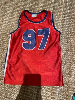 Röd linnetopp med blå detaljer Cinema - Sportig röd linnetopp från Cinema med blå kantband och stort tryck av siffran 97 i blått framtill. Perfekt för en cool och avslappnad stil. Materialet känns lätt och syntetiskt, vilket gör toppen luftig och bekväm.