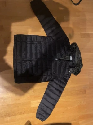 Svart pufferjacka från Stone Island - Säljer en svart pufferjacka från Stone Island med ikonisk patch på ärmen. Jackan har huva, dragkedja framtill och quiltad design. Perfekt för kalla dagar och riktigt snygg streetstil. Passar dig som vill ha en clean och modern look.