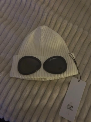 Vit ribbstickad mössa från C.P. Company - Säljer en vit ribbstickad mössa från C.P. Company med ikoniska svarta goggles framtill. Mössan har uppvikt kant och är tillverkad i mjukt, stretchigt material. Perfekt för dig som vill sticka ut med en streetwear-inspirerad look.
