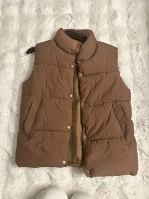 Brun dunväst från H&M - Snygg brun dunväst från H&M med hög krage och tryckknappar framtill. Västen har två fickor på sidorna och quiltad design som ger en trendig look. Perfekt att slänga över en hoodie för extra värme under kyliga dagar.
