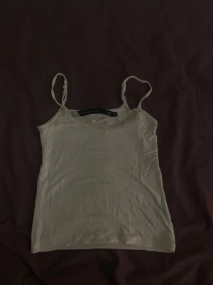 Vit linnetopp med spetsdetalj - Söt vit linnetopp från The Black Label Collection med tunna axelband och v-ringning dekorerad med spets. Perfekt att styla lager på lager eller bära som den är. Enkel och stilren design som passar till mycket.