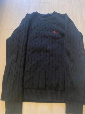 Svart kabelstickad tröja Polo Ralph Lauren - Kabelstickad svart tröja från Polo Ralph Lauren med klassisk röd logga broderad på bröstet. Tröjan har rund halsringning, ribbade muddar och är gjord i ett mjukt stickat material. Perfekt för dig som gillar stilrena och tidlösa plagg.