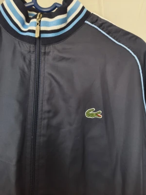 Marinblå ziptröja från Lacoste - Snygg marinblå ziptröja från Lacoste med klassisk krokodillogga på bröstet. Tröjan har vita och ljusblå ränder på krage, ärmslut och nederkant samt ljusblå piping längs ärmarna. Tillverkad i ett glansigt material som ger en sportig look.