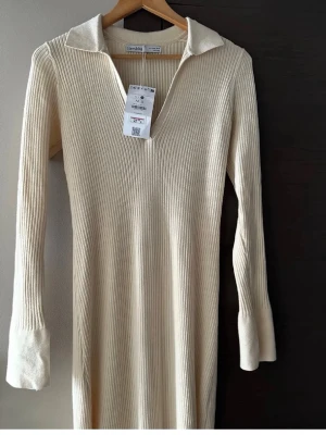Ribbad beige klänning från Bershka - Trendig ribbad klänning från Bershka i beige med v-ringning och krage. Klänningen har lång ärm och är tillverkad i ett mjukt, stickat material som ger en snygg och bekväm passform. Perfekt för dig som gillar stilrena och enkla plagg.