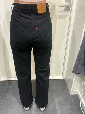 Svarta raka jeans från Levi's - Svarta jeans från Levi's med rak passform och klassiska fem fickor. Jeansen har hög midja, synliga nitar och Levi's ikoniska läderpatch bak i midjan. Materialet är kraftigt denim och bakfickorna har den typiska Levi's-sömmen.