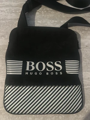 Svart axelväska från Hugo Boss - Snygg svart axelväska från Hugo Boss med vit logga och coola vita ränder på framsidan. Väskan har justerbar axelrem och dragkedja upptill. Tillverkad i slitstarkt tygmaterial och har en modern, fyrkantig form.