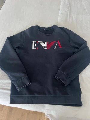 Mörkblå  sweatshirt från Emporio Armani - Snygg mörkblå sweatshirt från Emporio Armani med stort EVA-logotyptryck i vitt och rött på bröstet. Tröjan har rund halsringning, långa ärmar och ribbade muddar. Perfekt för en stilren och trendig look.