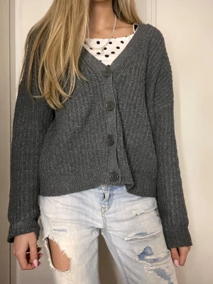 Mörkgrå stickad kofta med knappar - Mysig mörkgrå stickad kofta med v-ringning och stora knappar framtill. Koftan har en avslappnad passform och långa ärmar. Perfekt att slänga över en t-shirt eller linne för en chill look.