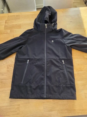 Svart vindjacka från 8848 Altitude - Svart vindjacka med huva från 8848 Altitude. Jackan har hel dragkedja framtill, två dragkedjeförsedda fickor och tryckt 8848-logga på ärmarna. Materialet är slätt och lätt, perfekt för blåsiga dagar. Knappt använd.