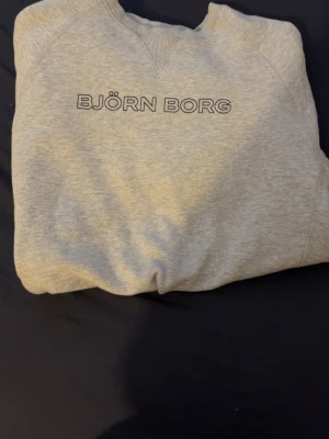 Grå sweatshirt från Björn Borg - Snygg grå sweatshirt från Björn Borg med klassisk logga tryckt på bröstet. Tröjan har rund halsringning med ribbade muddar och är tillverkad i mjukt material för extra komfort. Perfekt för en avslappnad och sportig stil.