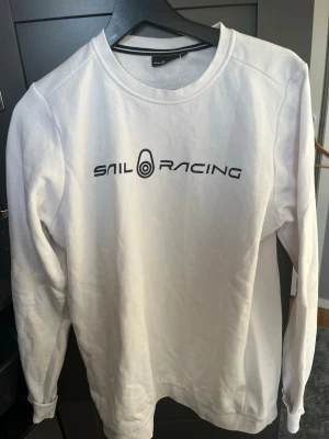Vit sweatshirt från Sail Racing - Vit sweatshirt från Sail Racing med svart logotyptryck på bröstet. Klassisk rund halsringning och långa ärmar. Tillverkad i mjukt material som känns skönt mot huden. Perfekt för en clean och sportig look.